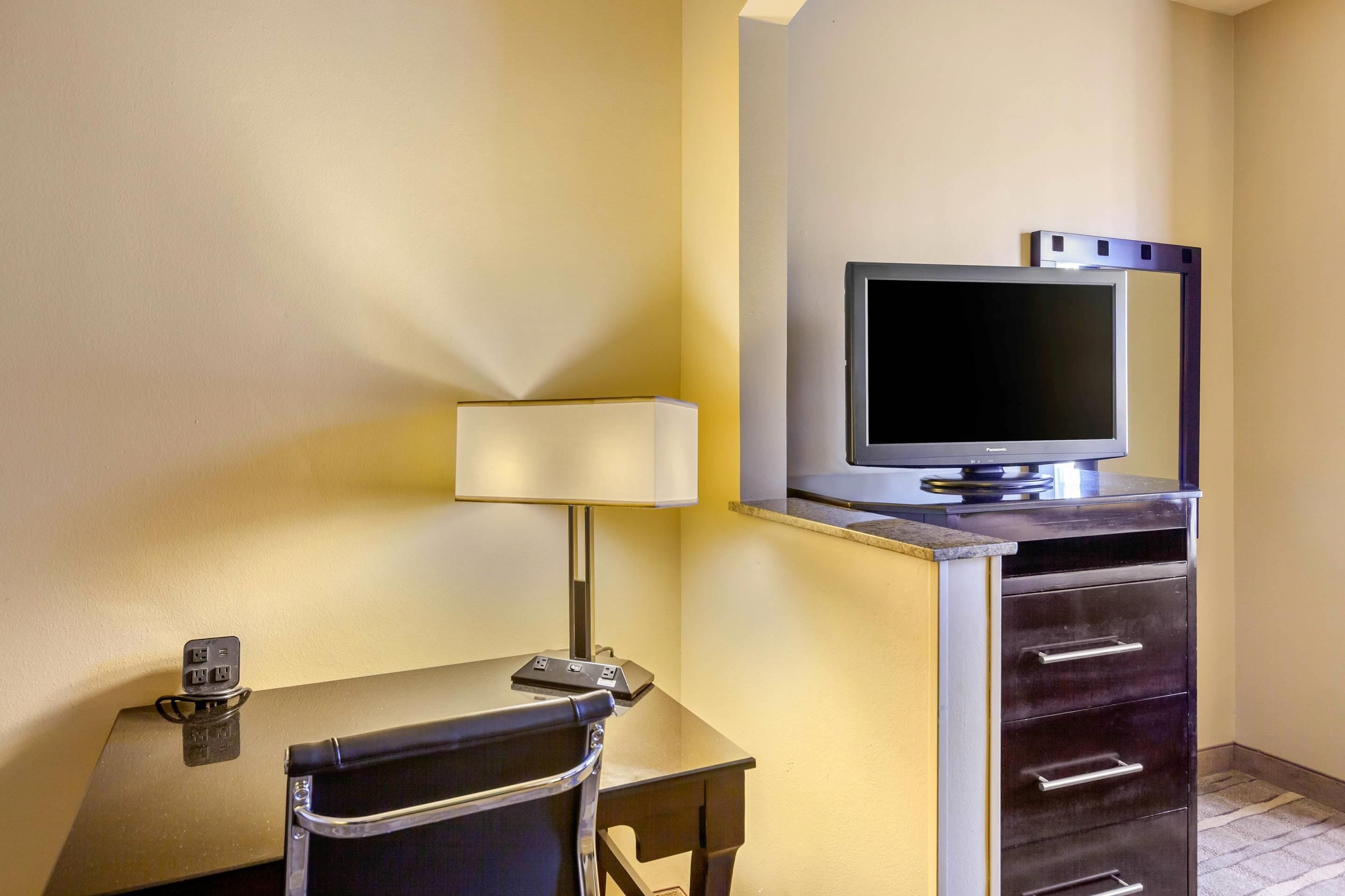 A hotel room with a desk, chair, lamp, television, and a minibar.