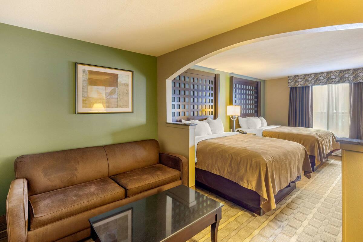 Habitación de hotel con sofá de cuero marrón, dos camas con ropa de cama marrón, un cuadro en la pared y una ventana con cortinas.