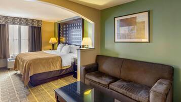 A hotel room with a bed, a sofa, a coffee table, a window with curtains, a wall-mounted lamp, and a framed picture.