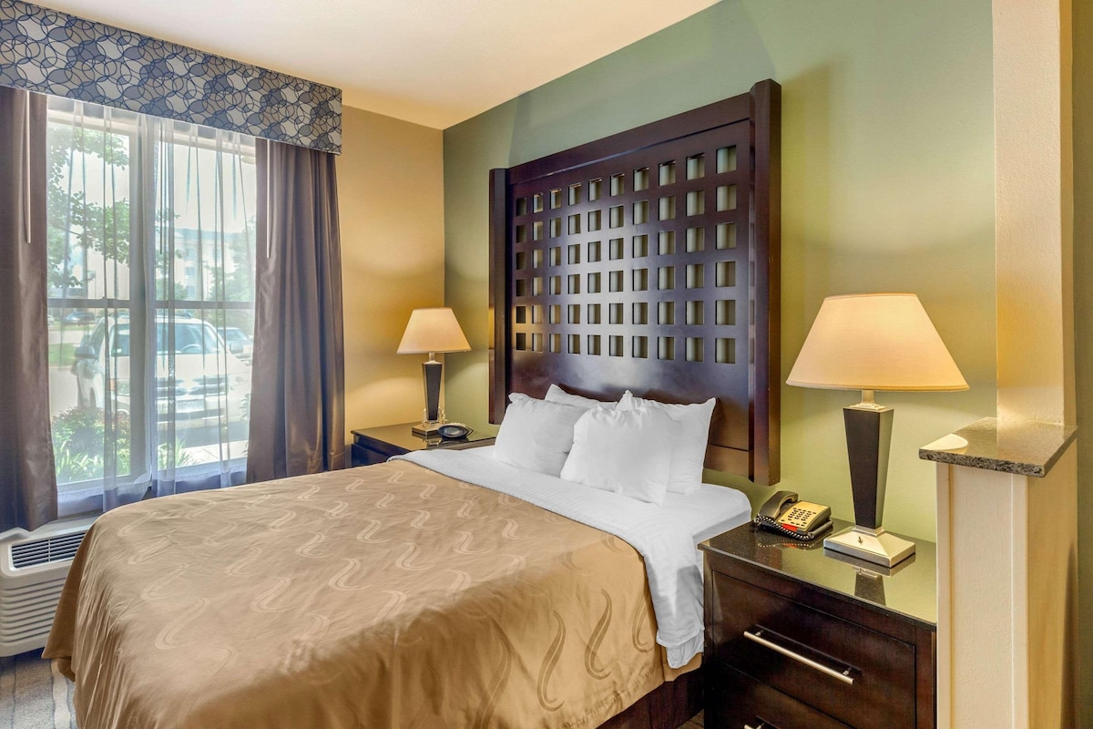 A hotel room with a large bed, two bedside tables with lamps, a wooden headboard, and a window with curtains.