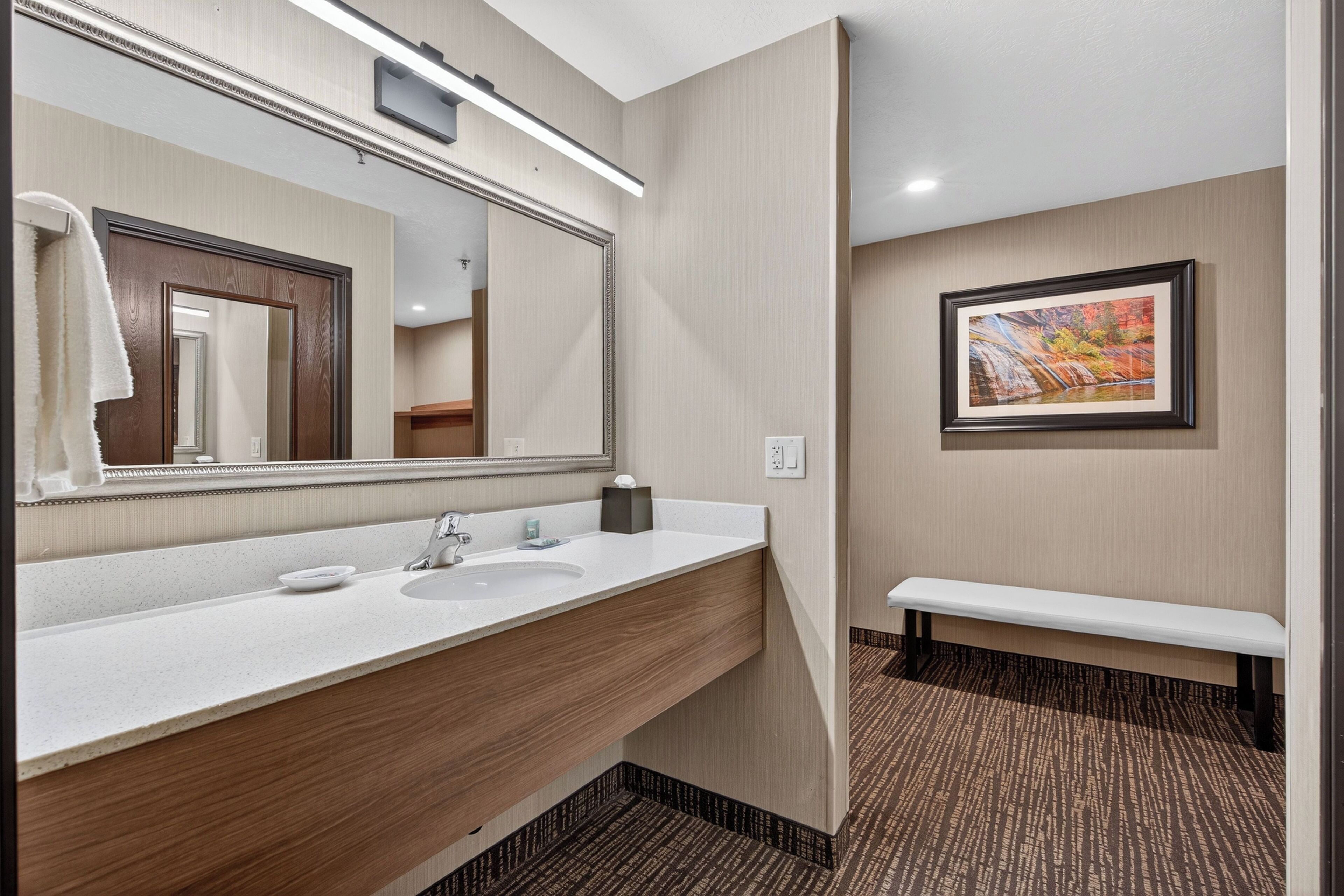 A hotel bathroom with a large mirror, a sink, a bench, and a framed picture on the wall.