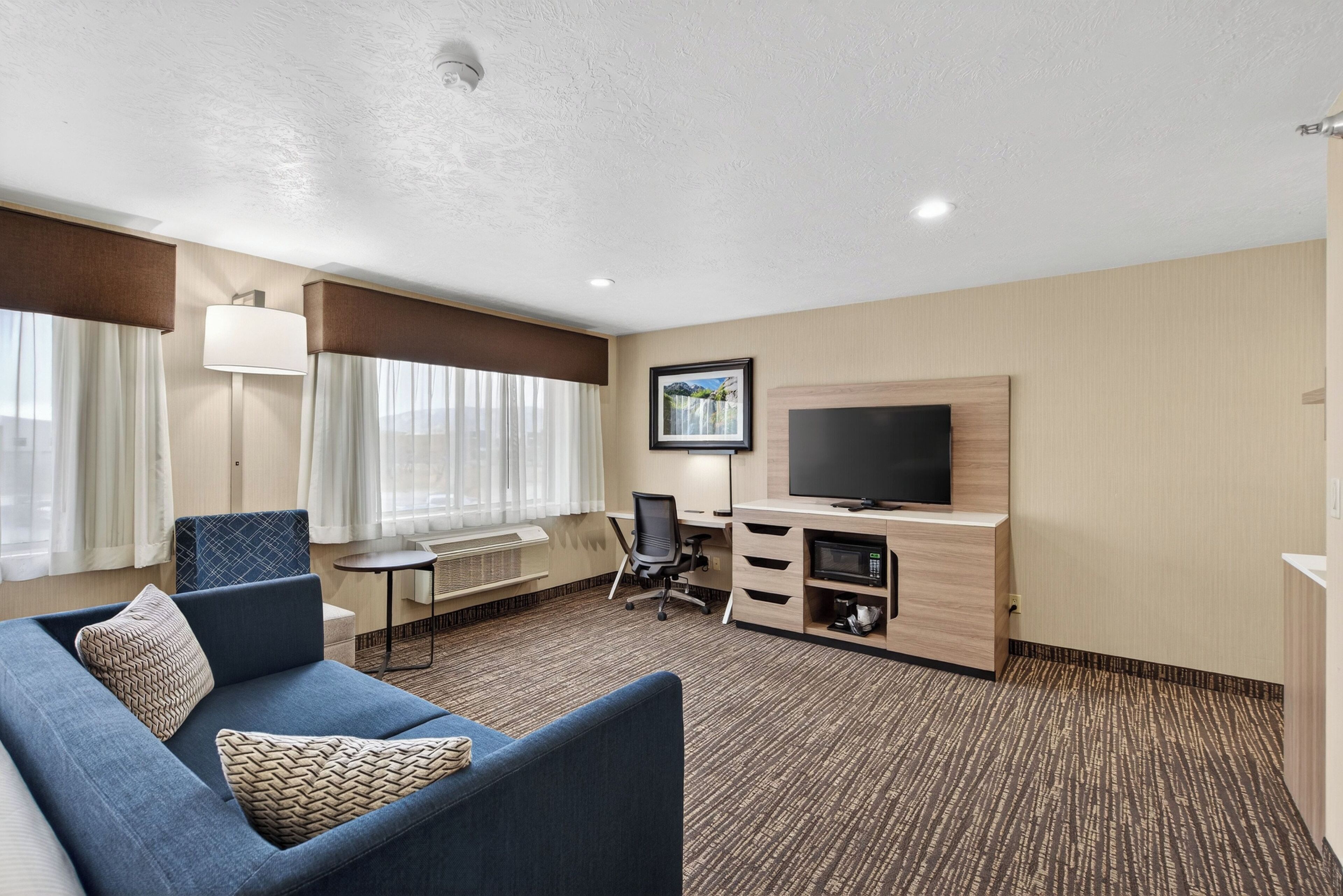 Une chambre d’hôtel avec un canapé bleu, un bureau avec un ordinateur, un téléviseur à écran plat et une fenêtre avec des rideaux.