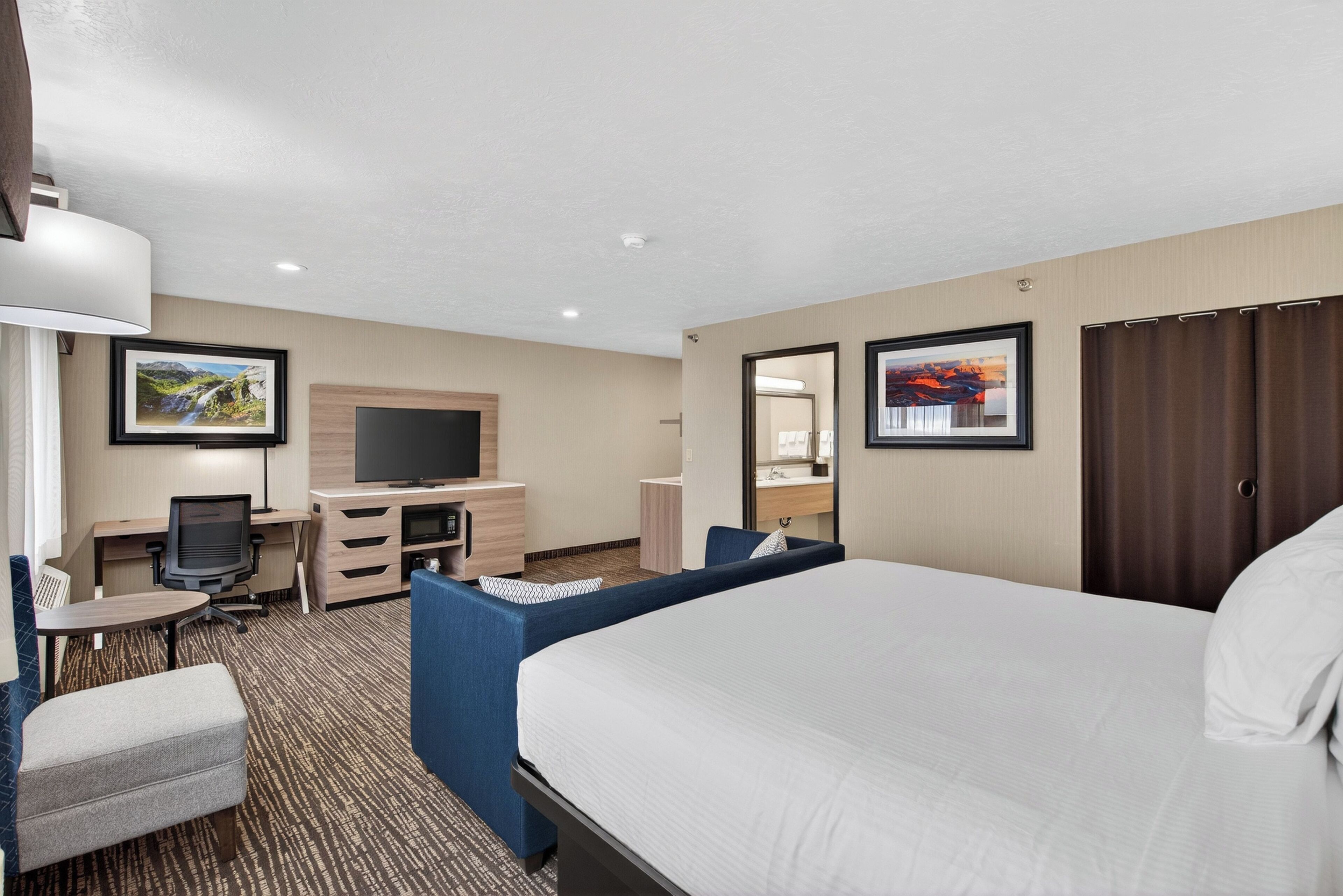 A hotel room with a large bed, a desk with a chair, a TV, and a small seating area.
