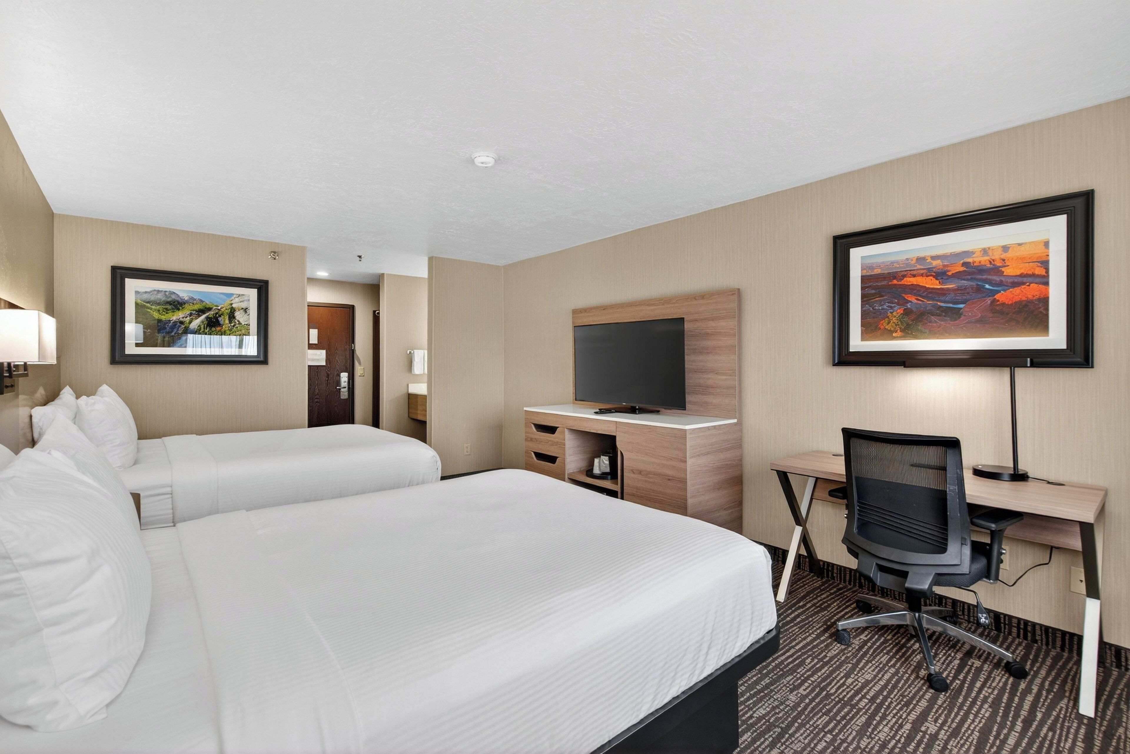 A hotel room with two beds, a desk with a computer, a TV, and framed pictures on the walls.
