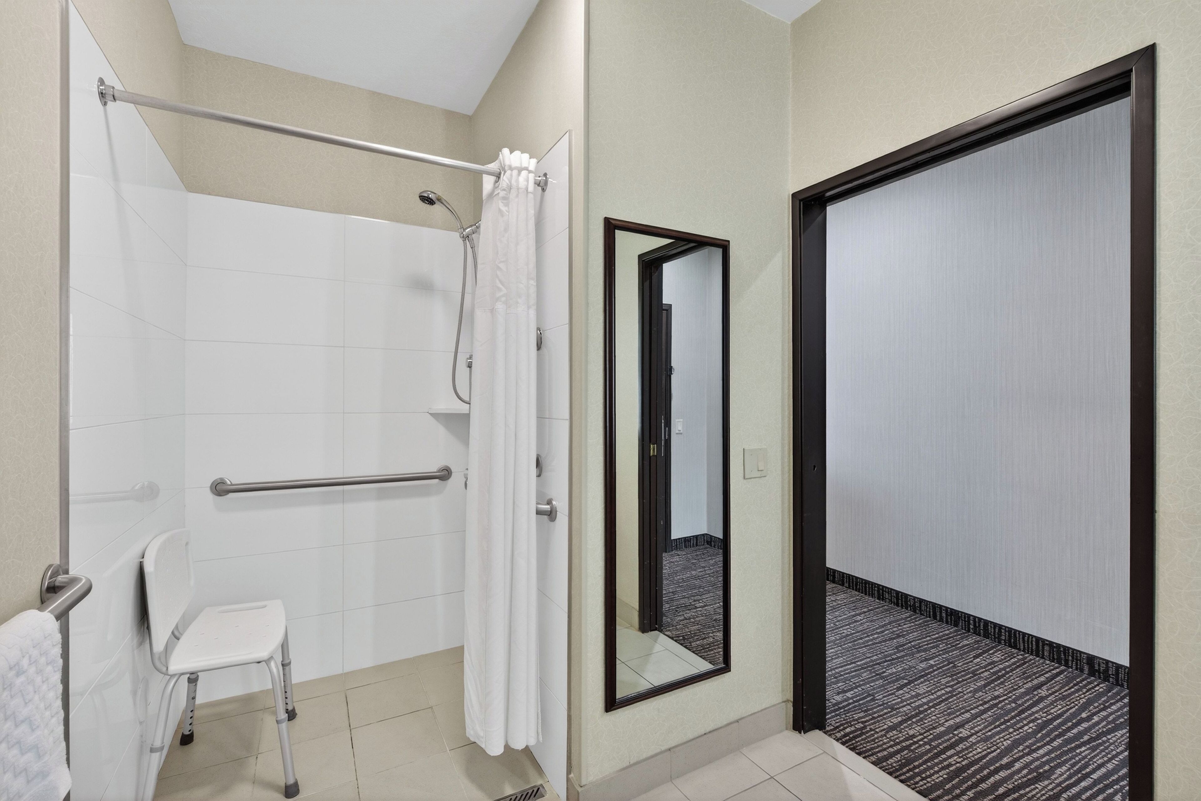 A modern bathroom with a shower, a white chair, a handrail, and a large mirror.