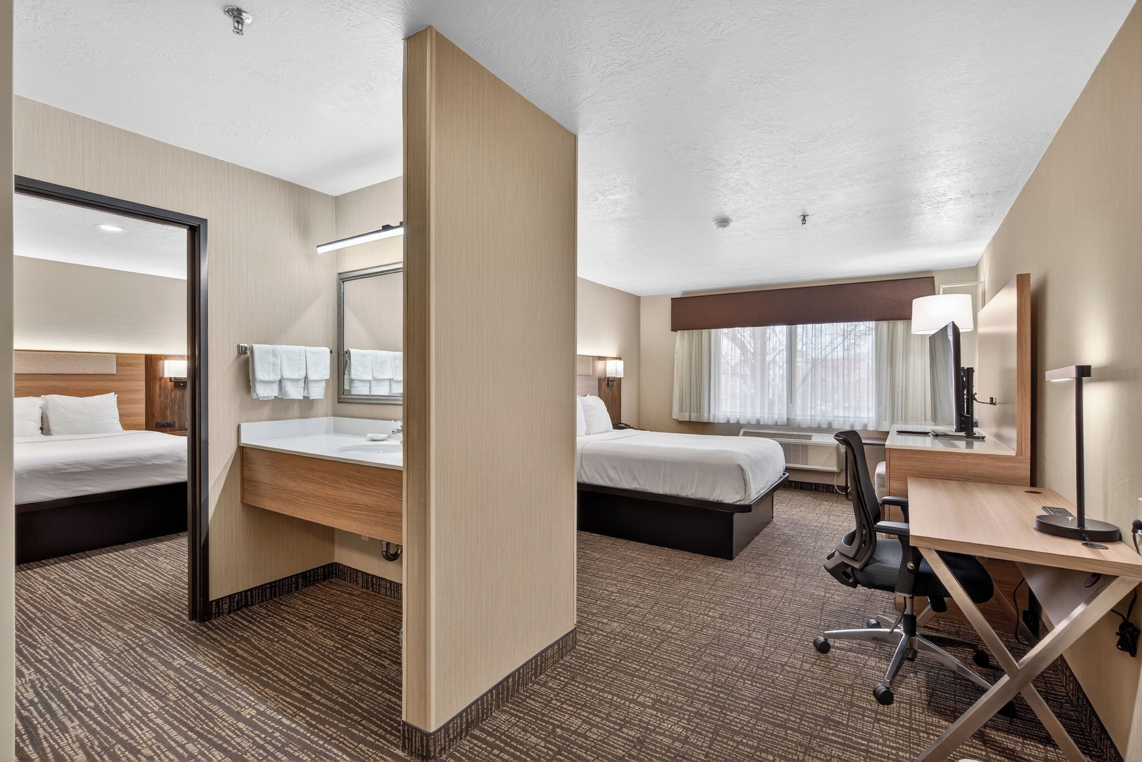 A hotel room with two beds, a desk, a chair, and a large window with curtains.