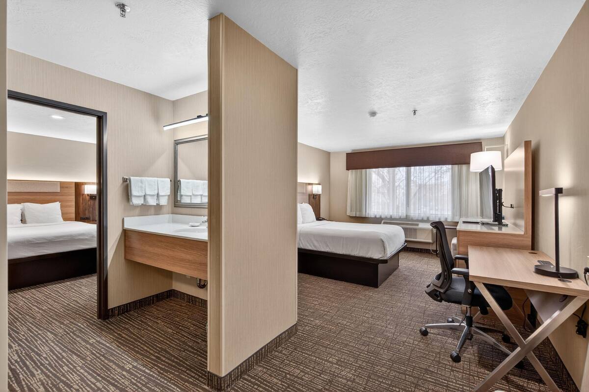 A hotel room with two beds, a desk, a chair, a bathroom, and a large window.