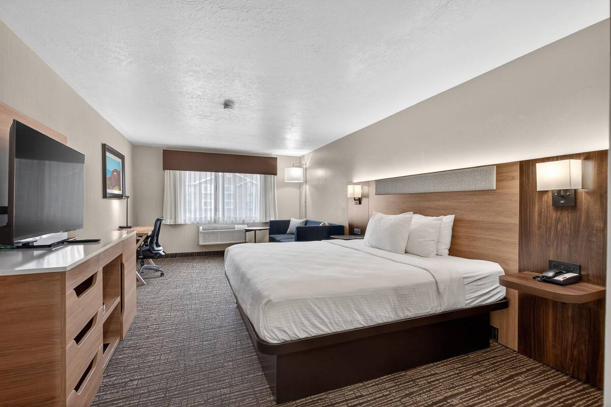 A hotel room with a large bed, a desk with a flat-screen TV, a chair, a lamp, and a small table.