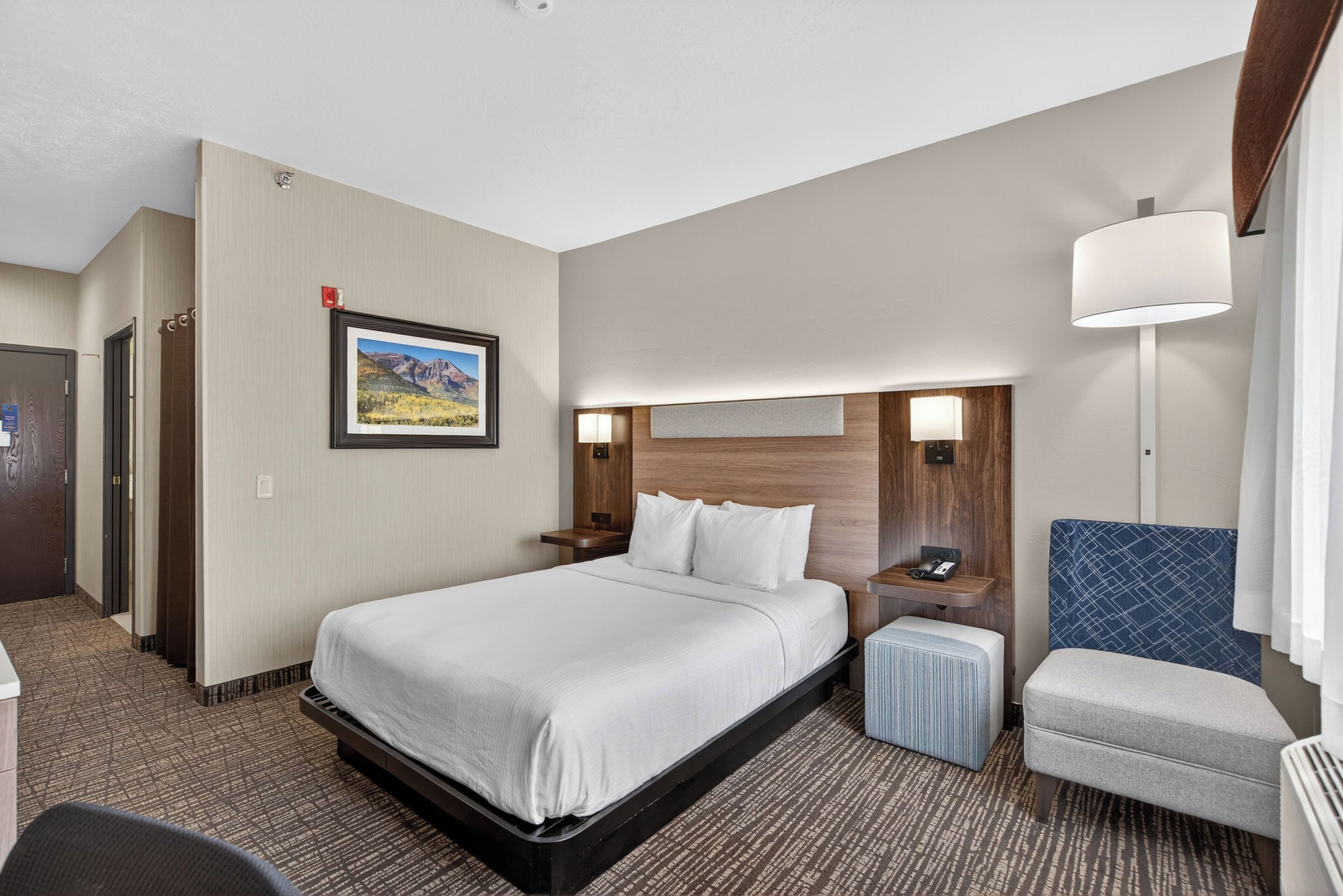 Habitación de hotel con una cama grande, un escritorio, una silla, una mesita, una lámpara y un cuadro en la pared.