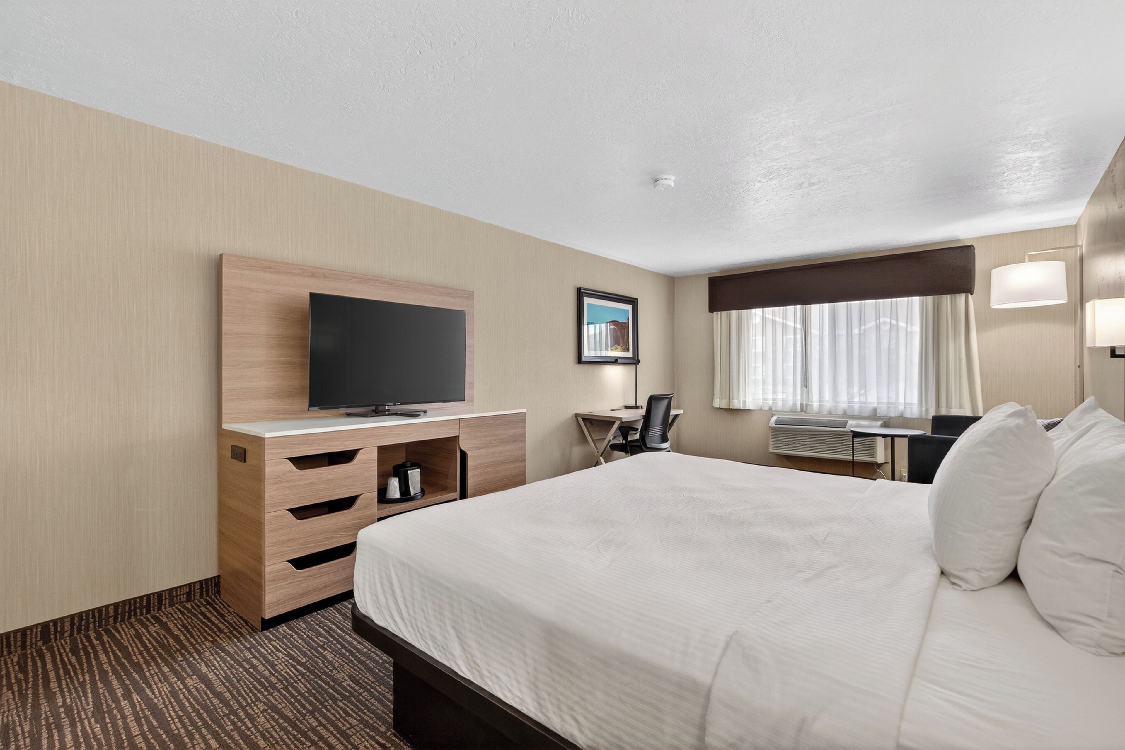 Quarto de hotel com uma cama grande, uma escrivaninha com cadeira, televisão de tela plana e uma janela com cortinas.