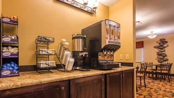 A hotel lobby with a coffee station, a buffet area with boxed food, and a dining area with a patterned carpet.