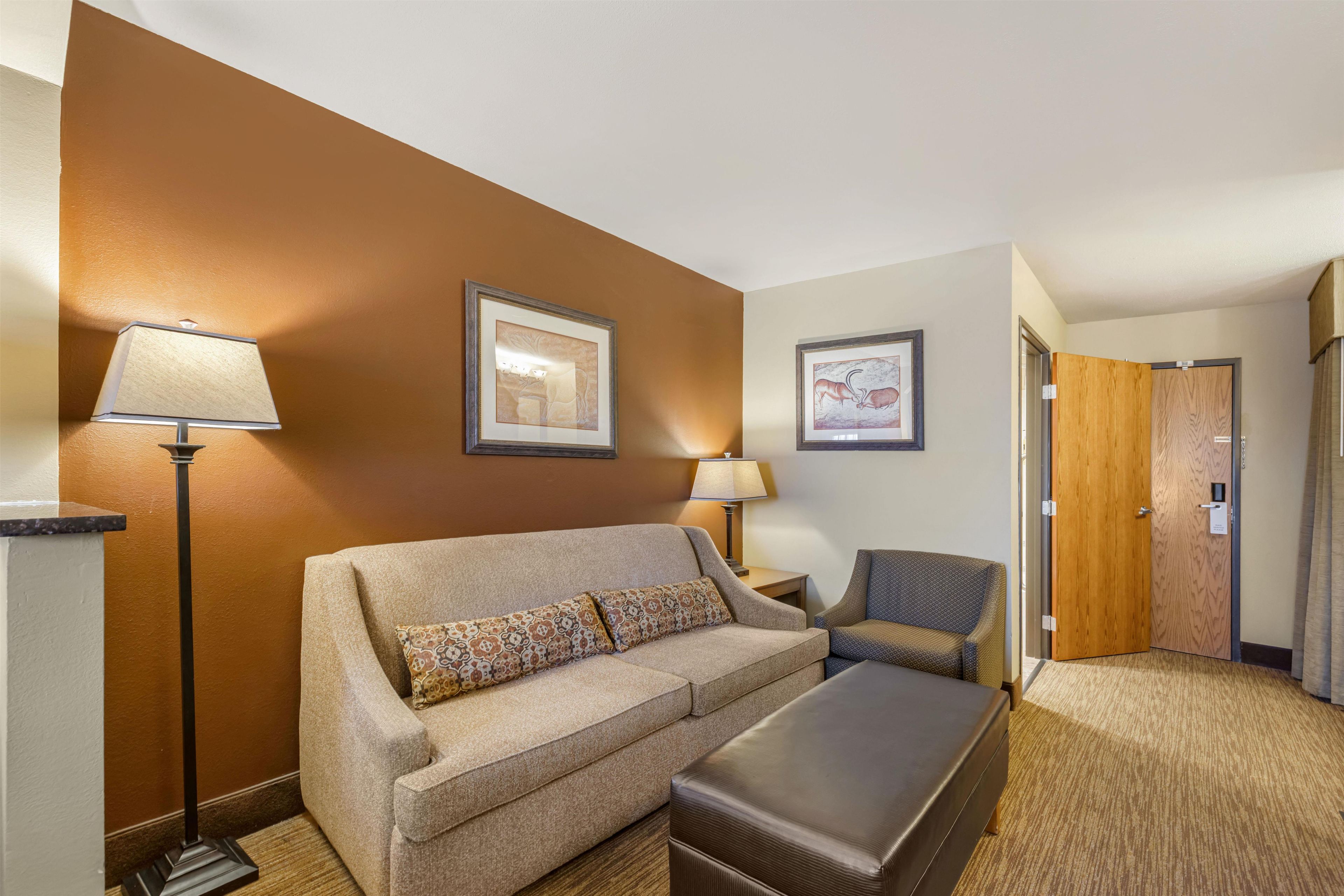 Une chambre d’hôtel avec un canapé, un fauteuil et une petite table. Il y a deux tableaux encadrés au mur, une lampe et une porte.