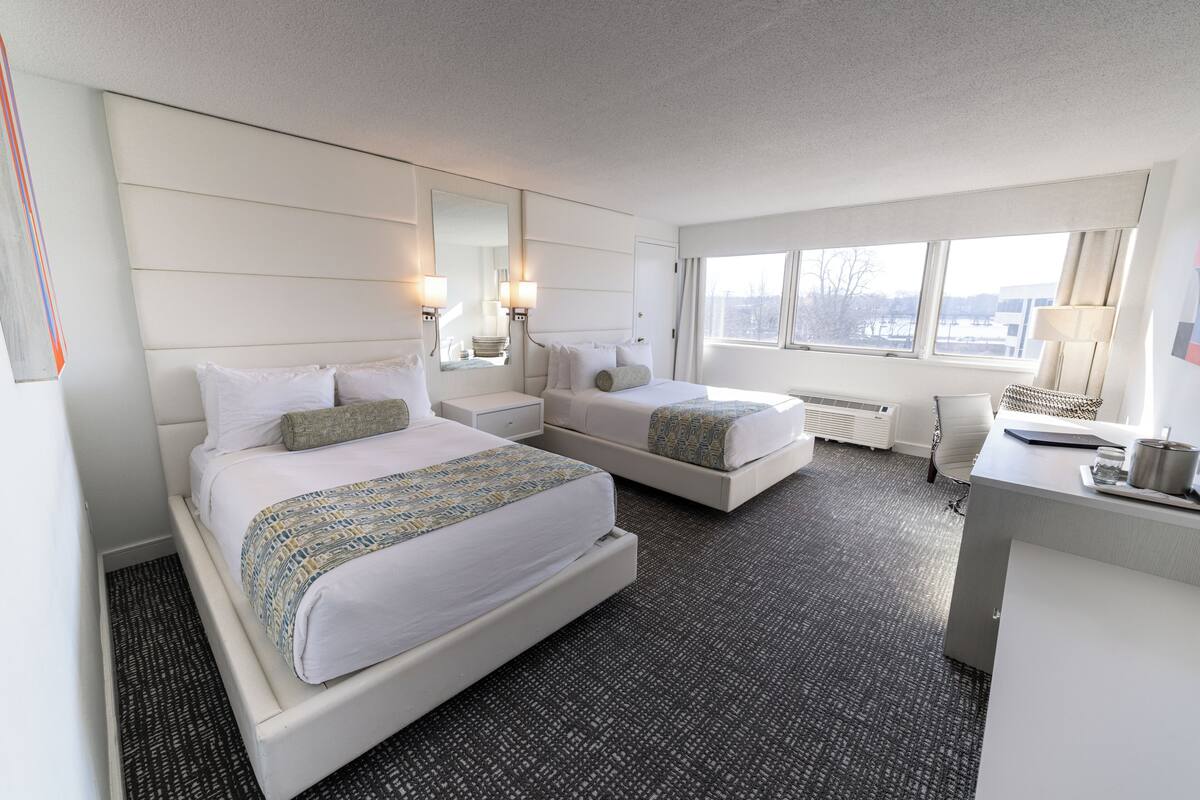 Ein Hotelzimmer mit zwei Betten, einem Schreibtisch, einem Stuhl und einem großen Fenster.
