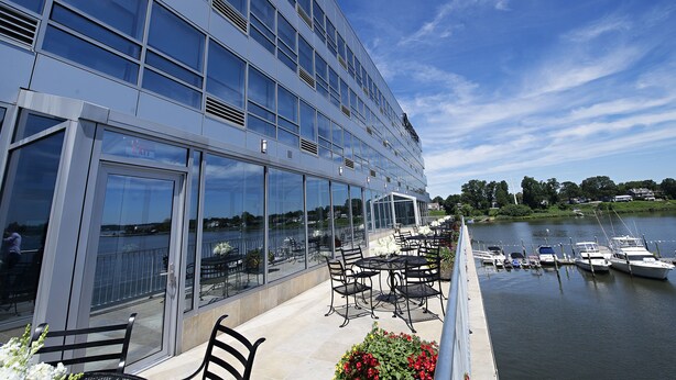 A modern building with large windows overlooking a calm body of water. There are outdoor seating areas with tables and chairs.