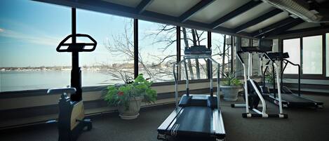 A gym with treadmills, an elliptical, and a stationary bike, large windows with a view of a body of water and trees.