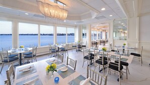 A dining area with tables set for service, large windows overlooking a body of water, and a chandelier hanging from the ceiling.