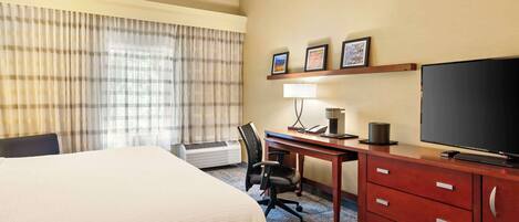 A hotel room with a large bed, a desk with a computer, a flat-screen TV, and a wall-mounted shelf with pictures.