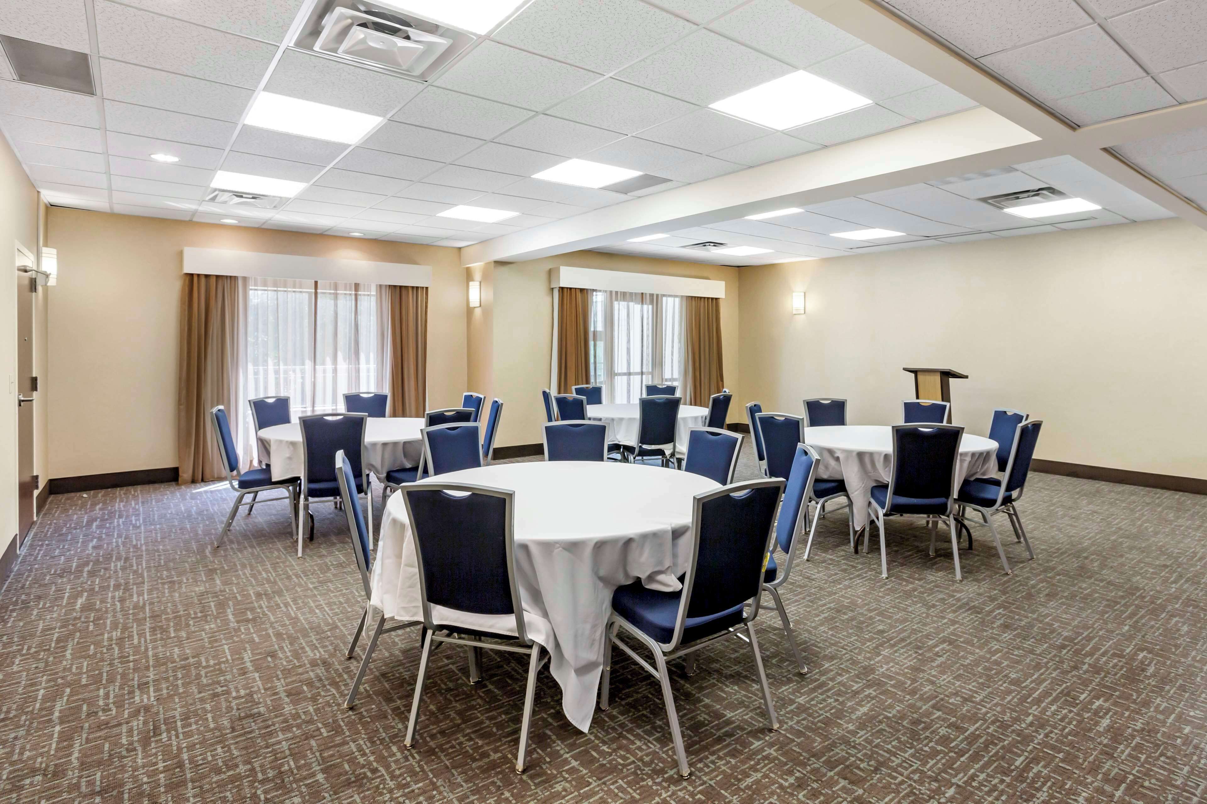A conference room with multiple round tables and chairs, large windows with curtains, and a carpeted floor.
