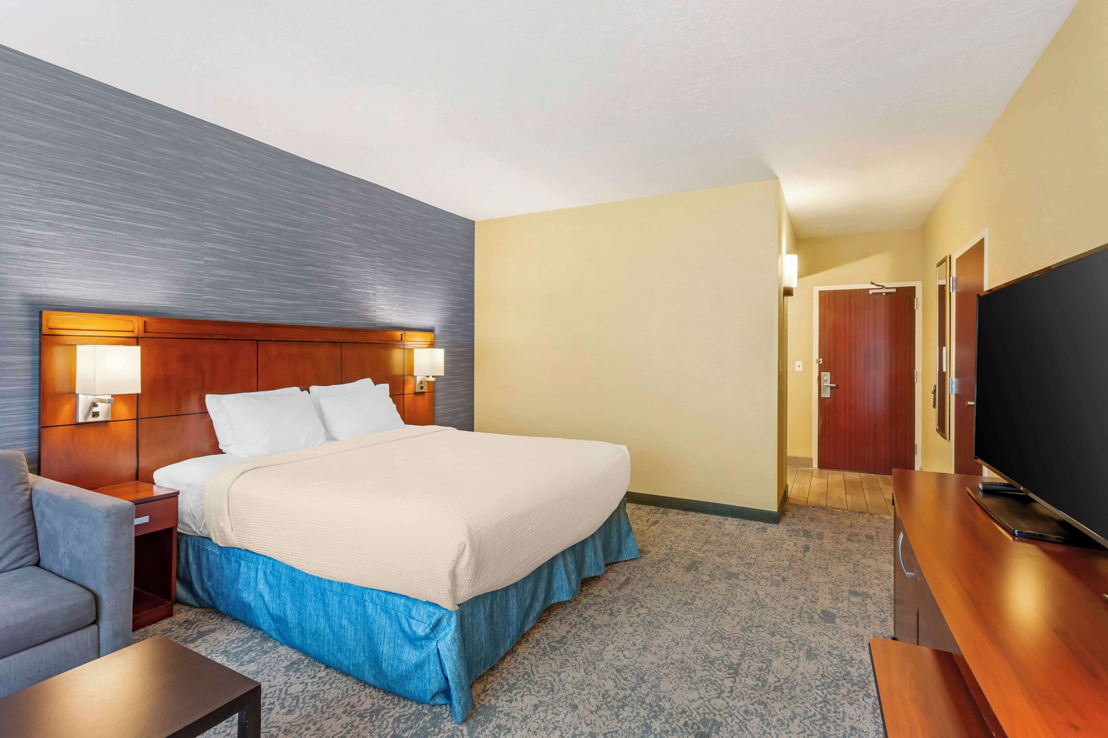 A hotel room with a bed, bedside table, lamp, TV, and a desk with a chair.
