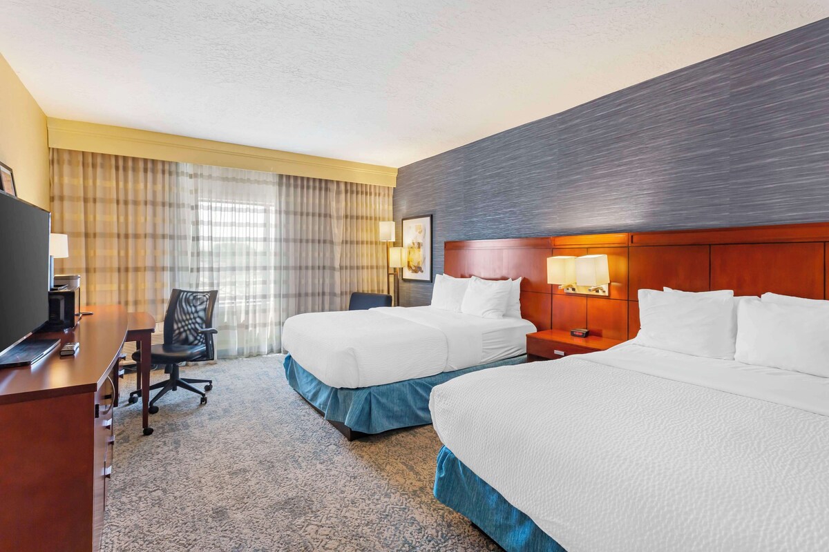 A hotel room with two beds, a desk, a chair, a TV, and a window with curtains.