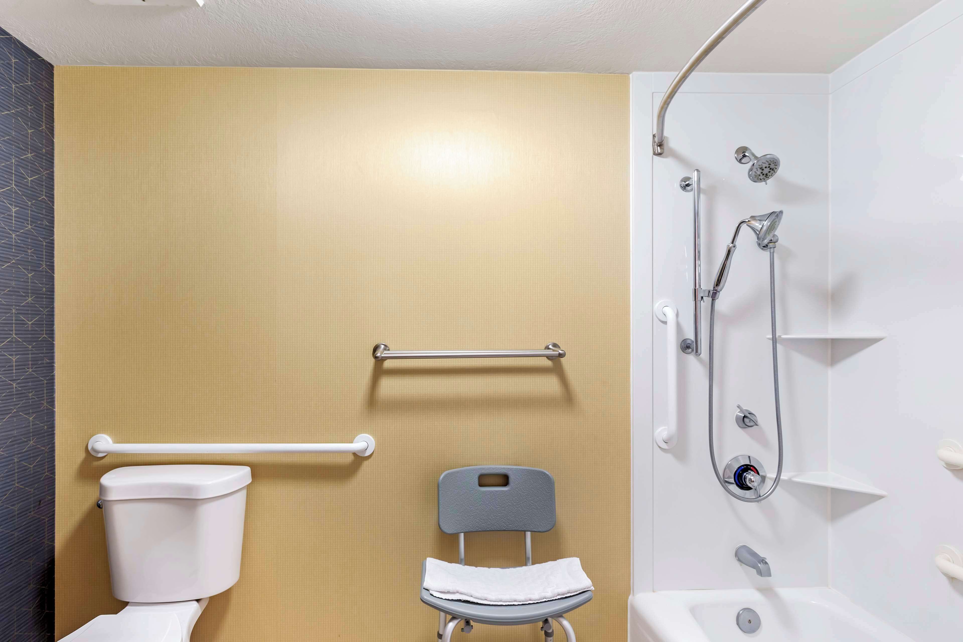 A bathroom with a toilet, a shower with a handheld showerhead, a grab bar, and a raised toilet seat.