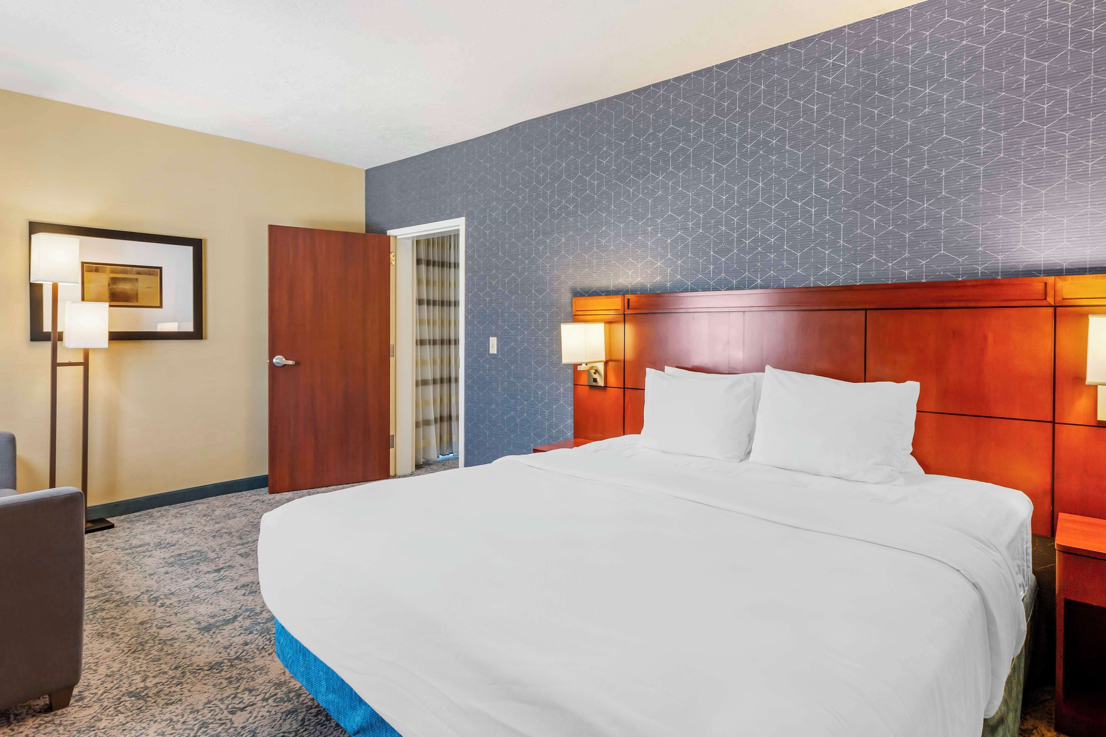 A hotel room with a large bed, a wooden headboard, two bedside lamps, a framed picture on the wall, and a carpeted floor.