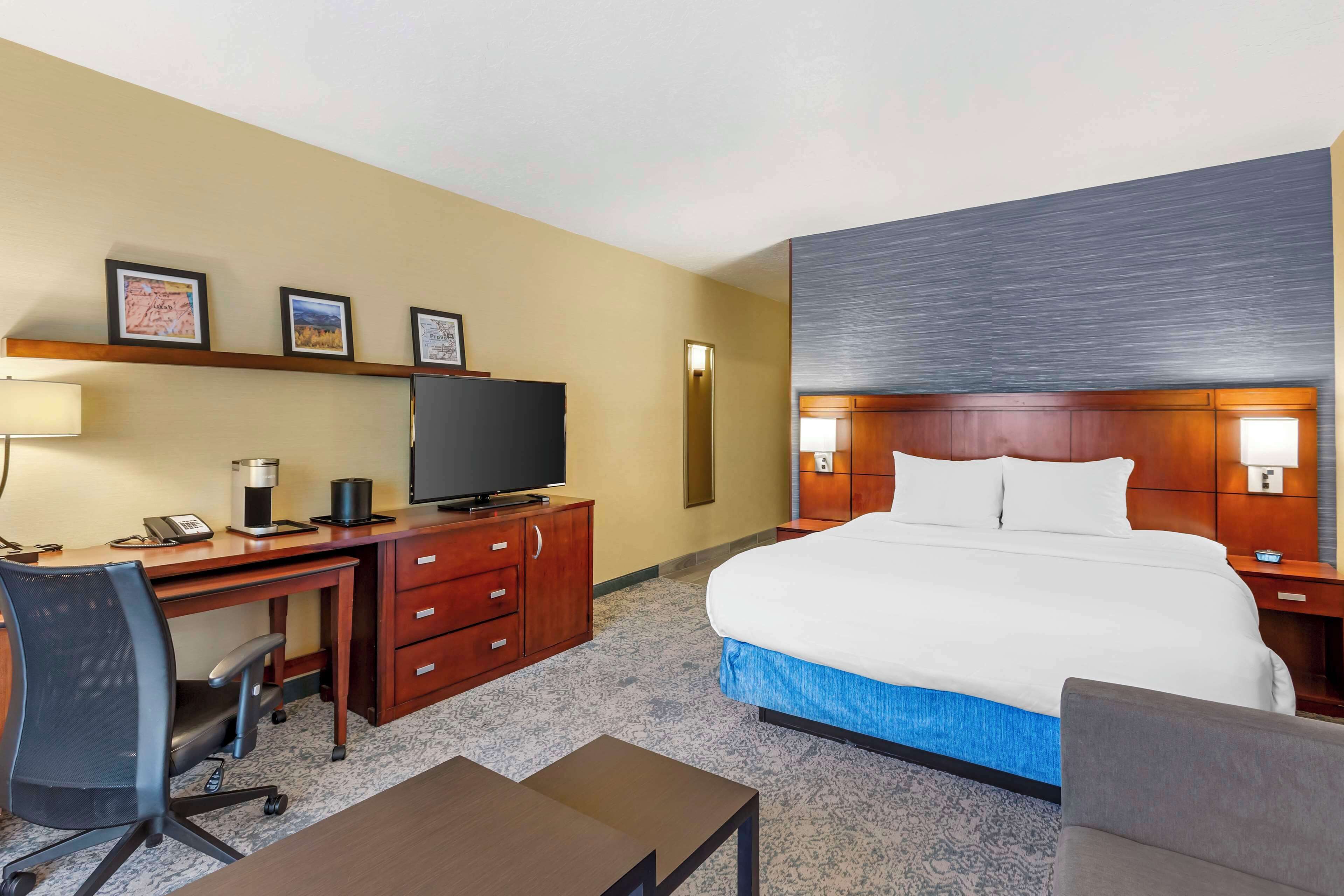 A hotel room with a large bed, a desk with a computer and a TV, a chair, a small table, and a nightstand with a lamp.