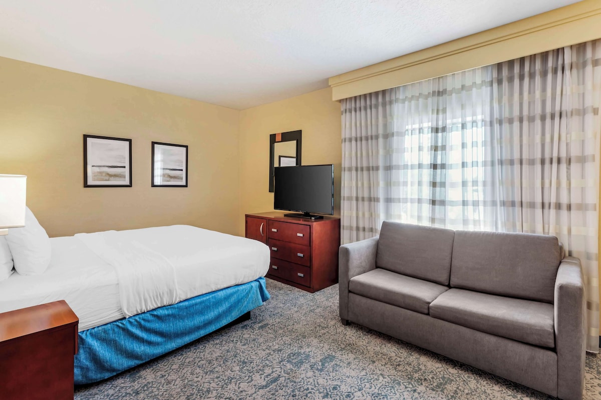 A hotel room with a bed, a sofa, a TV on a wooden cabinet, and two framed pictures on the wall.