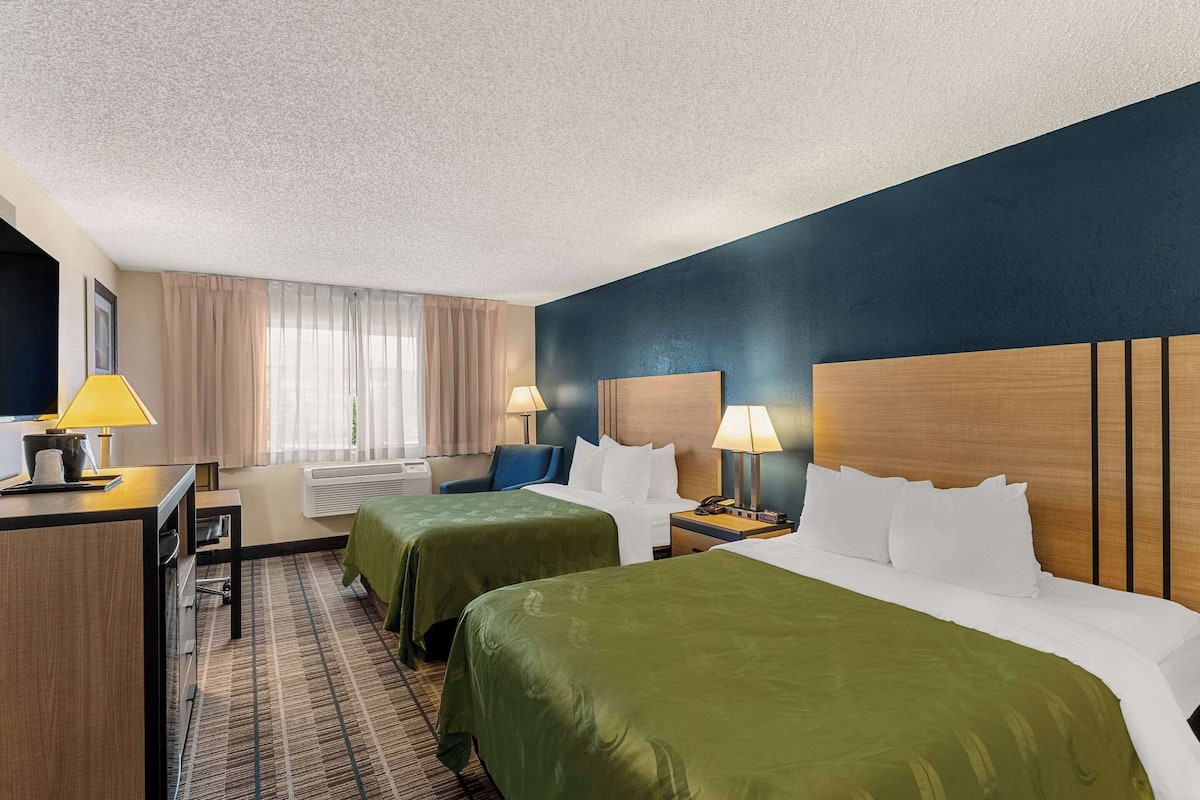 A hotel room with two beds, a desk, and a chair. There are lamps on the desk and nightstands beside the beds.