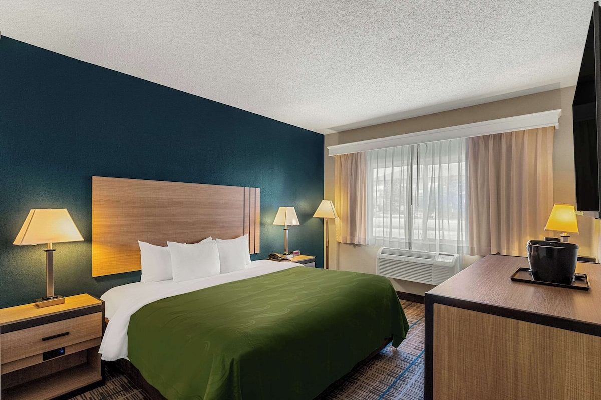 A hotel room with a large bed, two bedside tables with lamps, a desk with a lamp, a TV, and a window with curtains.