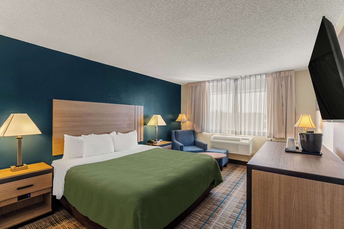 A hotel room with a large bed, two bedside tables with lamps, a desk with a flat-screen TV, a chair, and a window with curtains.