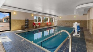 Una piscina interior con agua cristalina, un trampolín y una zona de estar con sillas rojas.