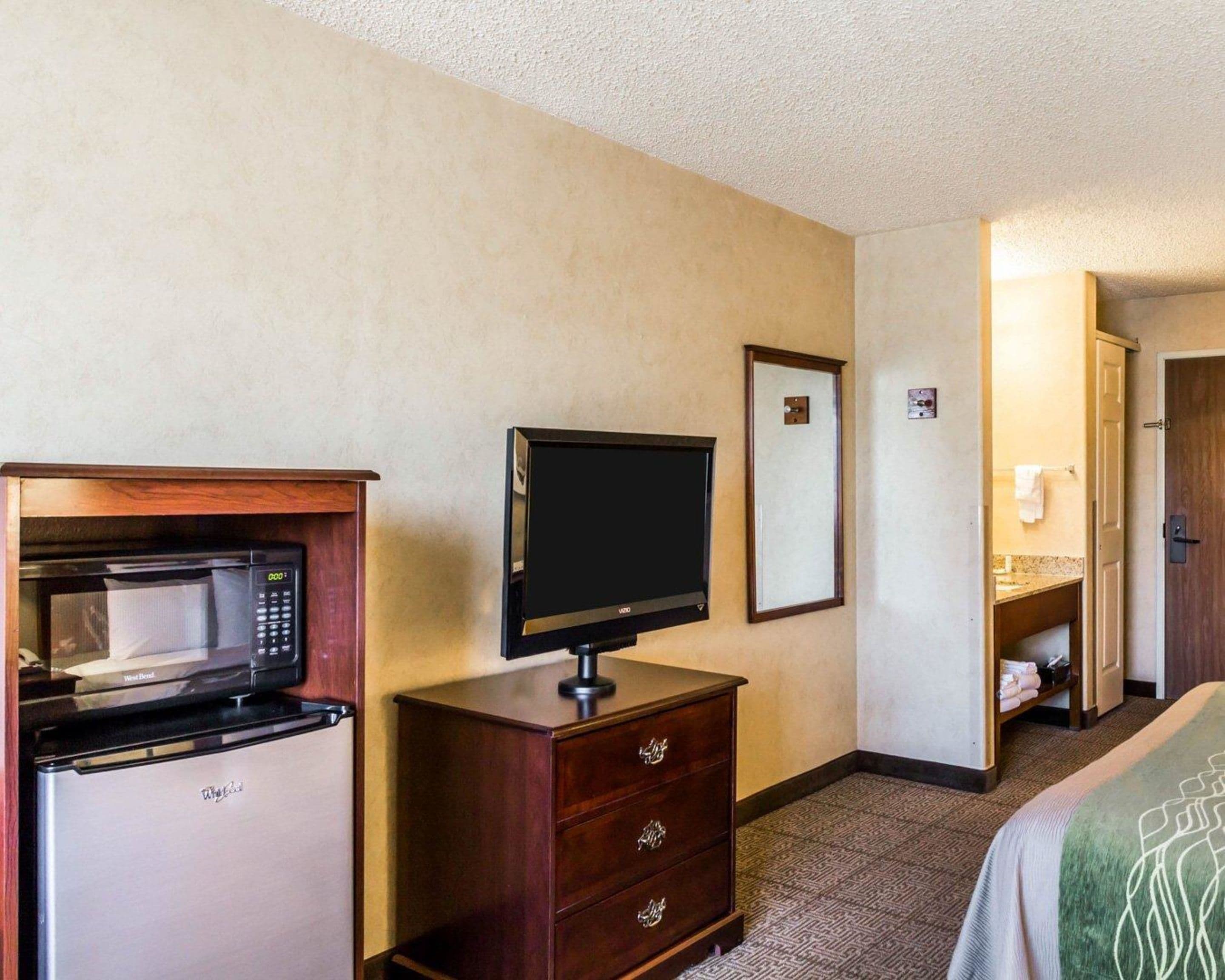 A hotel room with a kitchenette including a microwave, a flat-screen TV, a wooden dresser, and a bathroom with a vanity and towel rack.