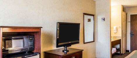 A hotel room with a kitchenette including a microwave, a flat-screen TV, a wooden dresser, and a bathroom with a vanity and towel rack.