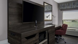 A modern hotel room with a large flat-screen TV mounted on a dark wood cabinet, a red office chair, a microwave, and a large window with a view of the outdoors. - Hampton Inn Conyers (Conyers)