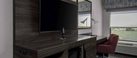 Habitación de hotel moderna con un televisor de pantalla plana grande montado en un mueble de madera oscura, una silla de oficina roja, un microondas y un amplio ventanal con vistas al exterior.