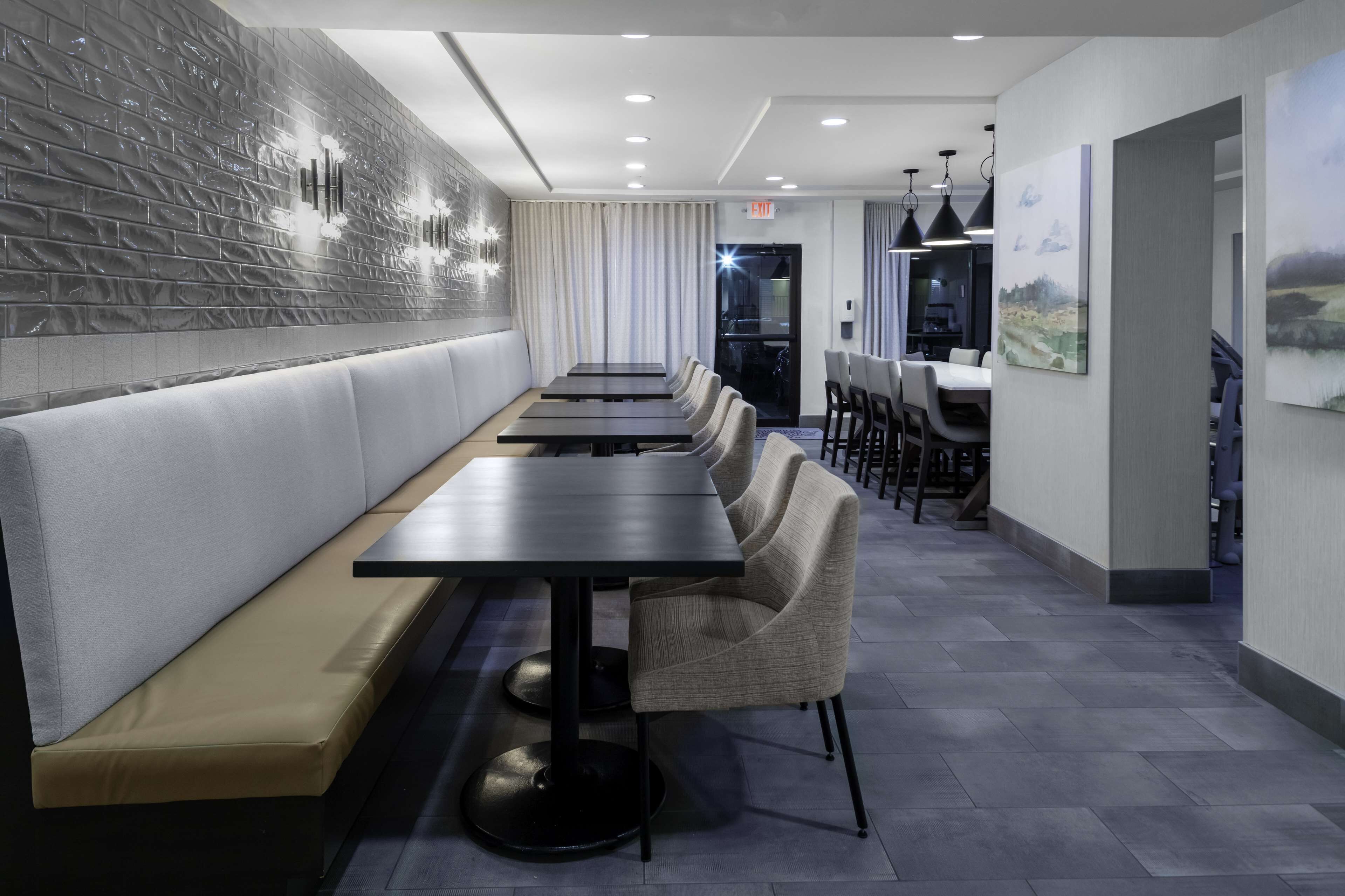 Un restaurante moderno con un largo banco y varias mesas, una pared de ladrillo con luminarias y vista a otra sala.