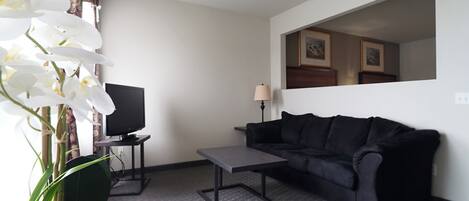 A living room with a black sofa, a small table, a TV on a stand, a potted orchid, and a lamp.