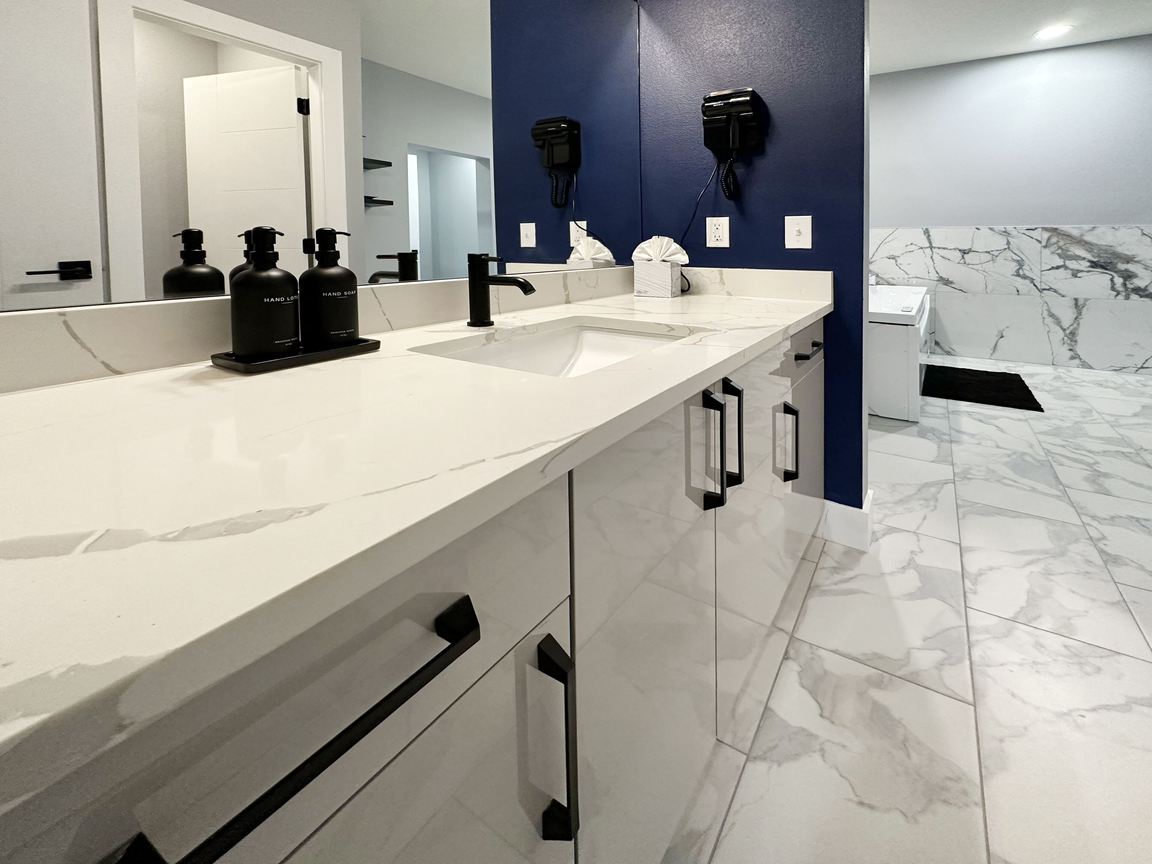 A modern bathroom with a white marble sink, black soap dispensers, a mirror, and a blue accent wall.