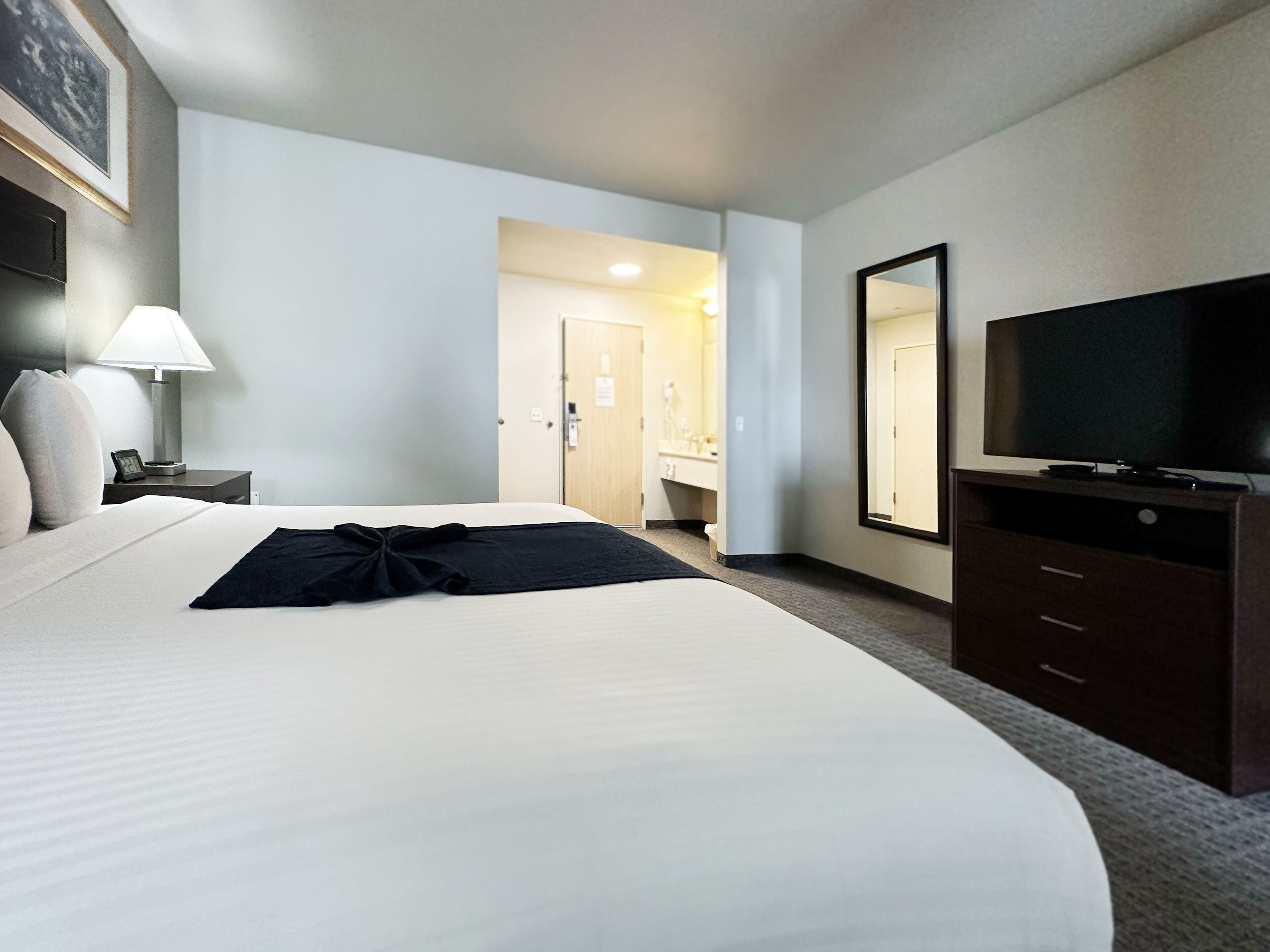 A hotel room with a large bed, a TV, a dresser, a nightstand with a lamp, and a door leading to a bathroom.
