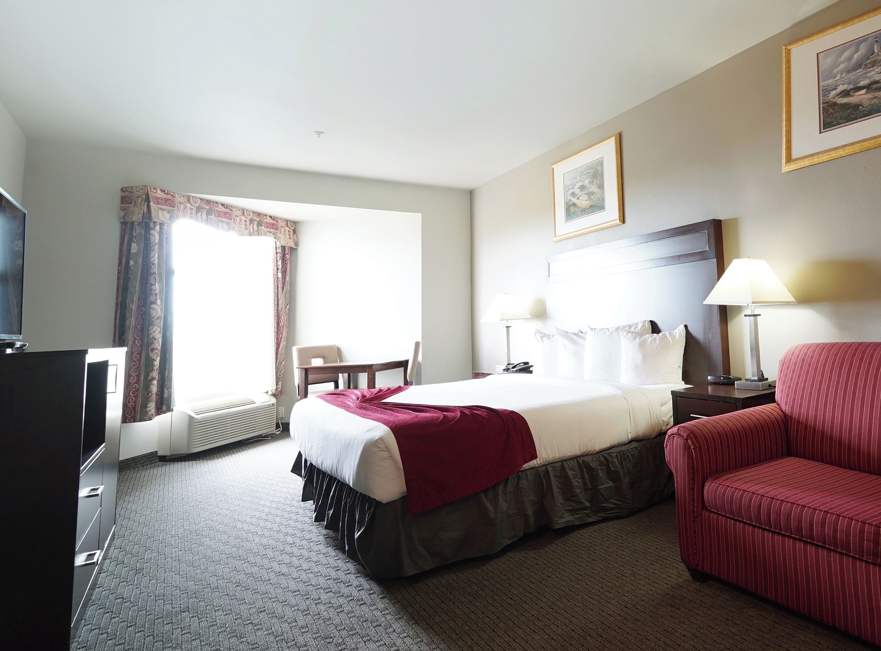A hotel room with a bed, a red sofa, a desk with a lamp, a chair, and a television.