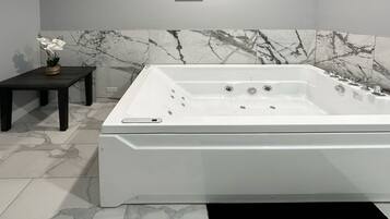 Un baño moderno con una bañera blanca grande, paredes de mármol y una mesita con una planta en maceta.