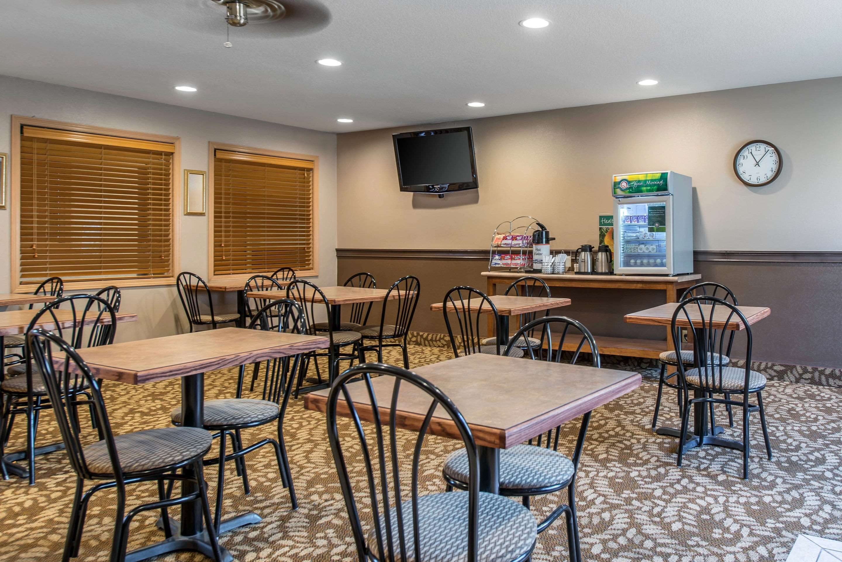 A hotel dining area with multiple tables and chairs, a television mounted on the wall, a clock, and a coffee machine.