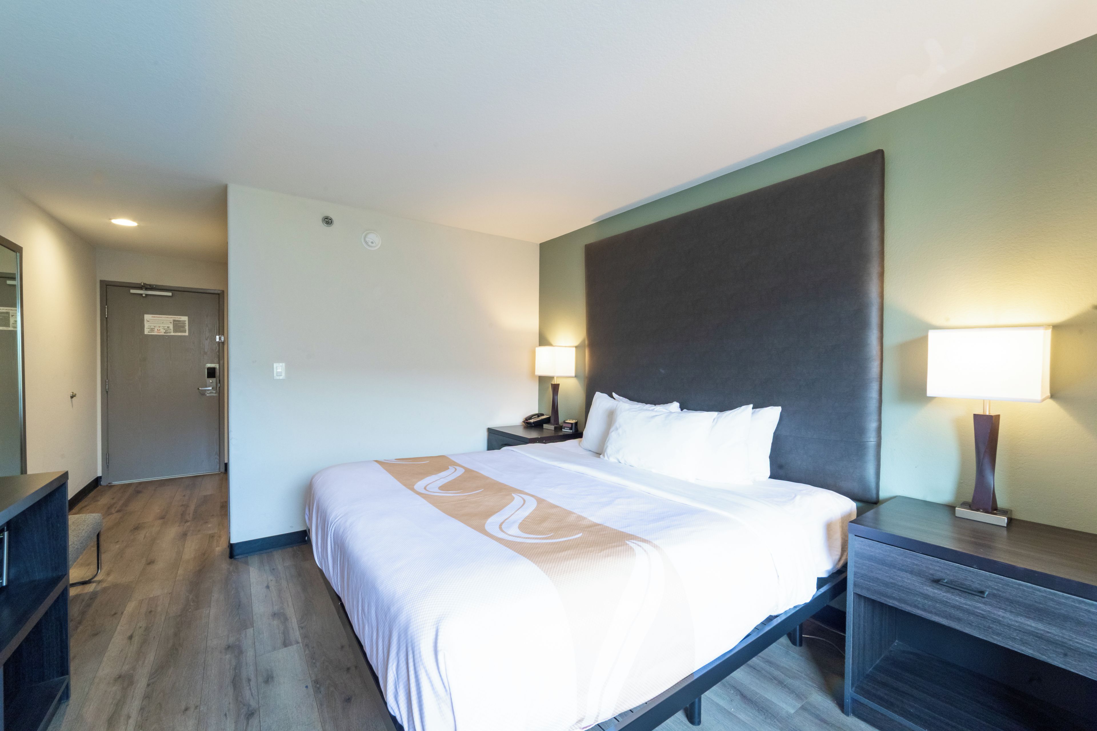 A hotel room with a large bed, bedside tables, a desk, and a door.