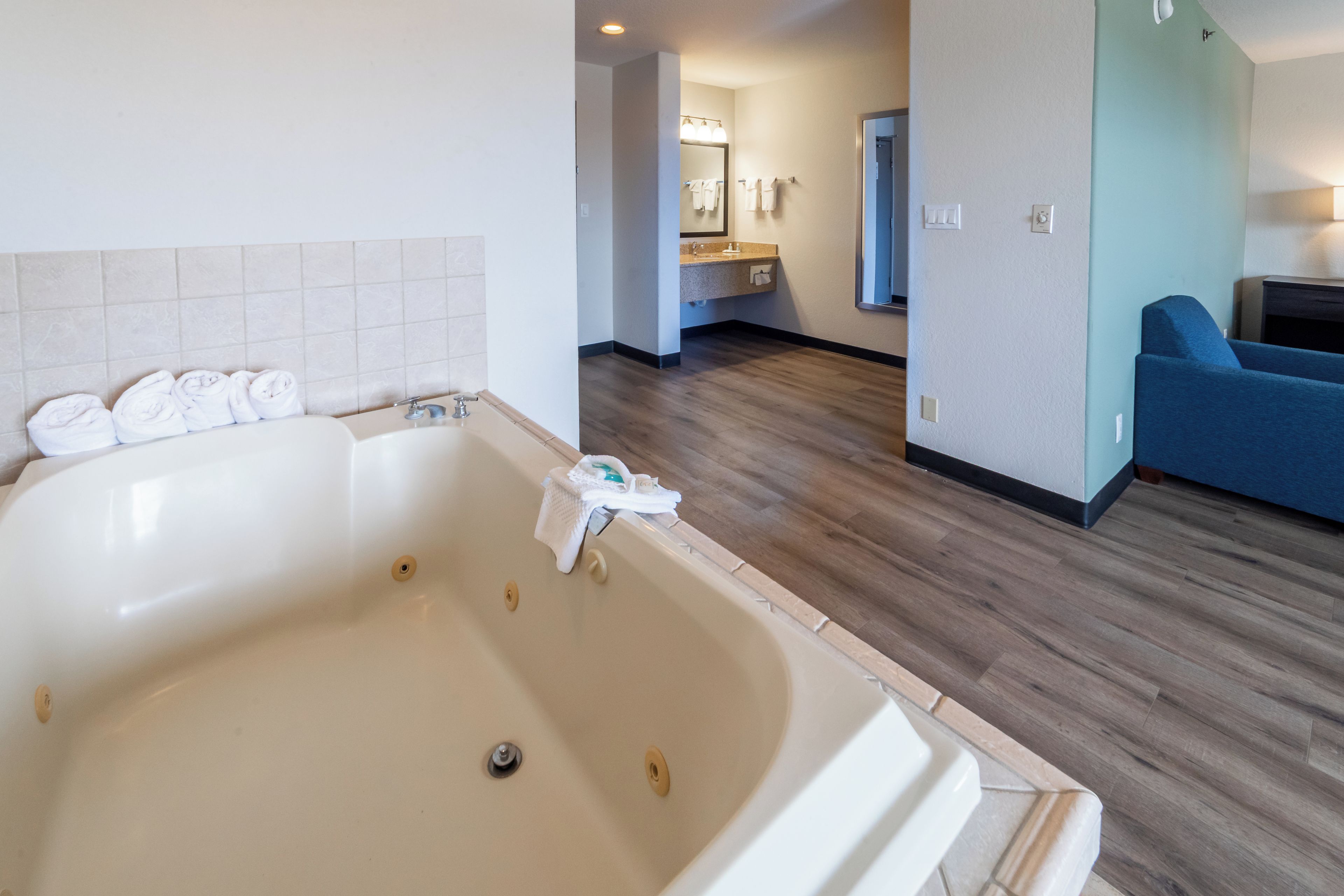 A hotel room with a large bathtub, a wooden floor, a blue sofa, and a bathroom with a sink and mirror.