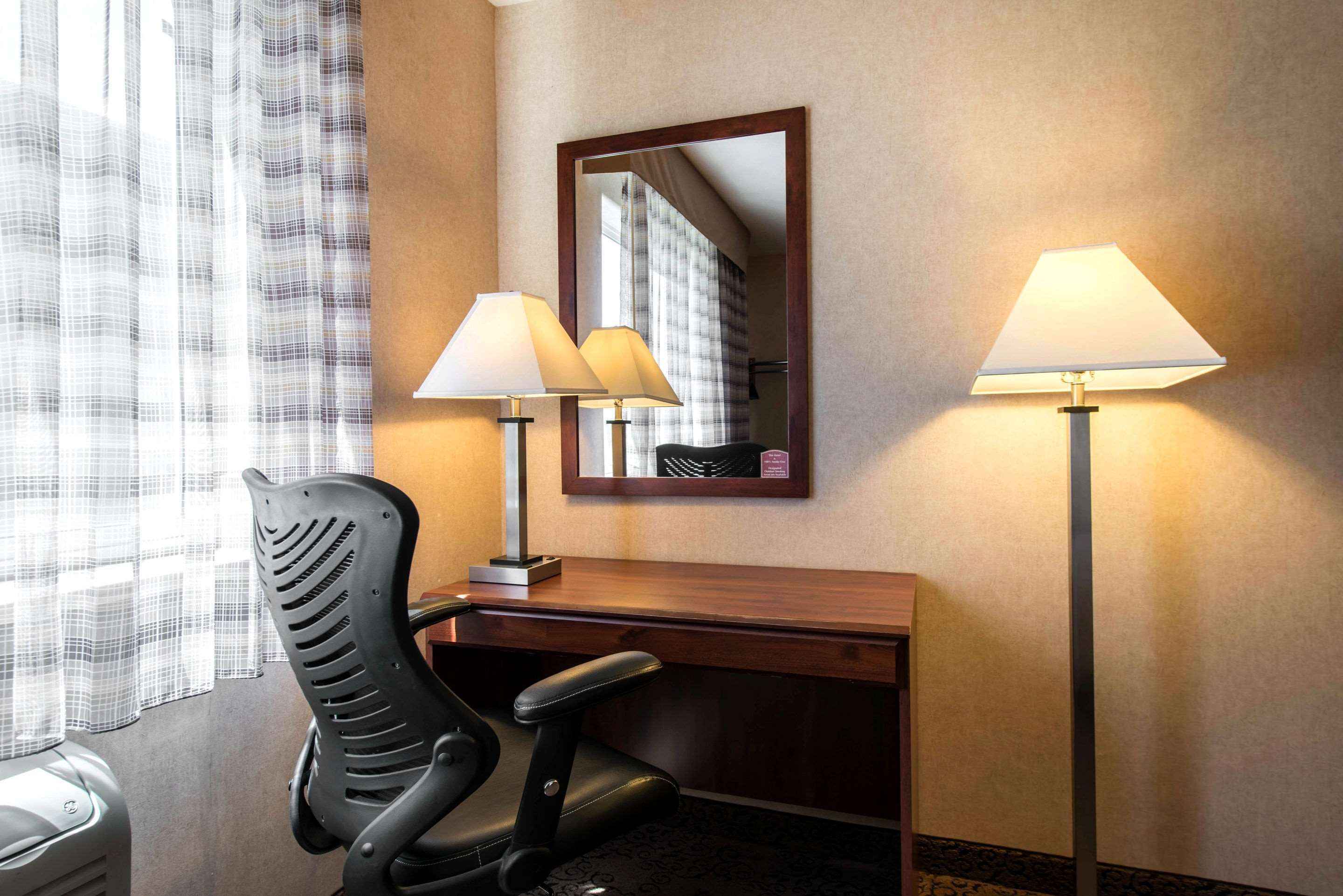 A hotel room with a desk, two lamps, a chair, and a mirror.