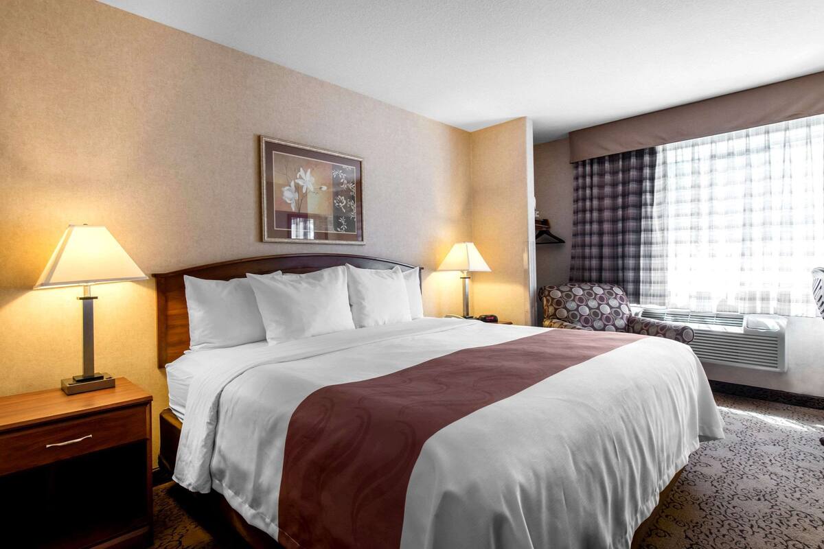 A hotel room with a large bed, two bedside lamps, a framed artwork on the wall, and a window with curtains.
