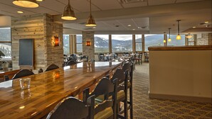 Un long comptoir en bois, agrémenté de nombreuses chaises, de luminaires suspendus et d’un mur en pierre avec une cheminée. De grandes fenêtres offrent une vue sur les montagnes.