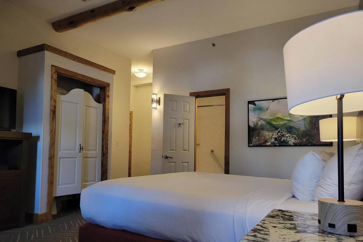 Een hotelkamer met een groot bed, een nachtkastje met lamp, een schilderij aan de muur en een houten balk in het plafond.