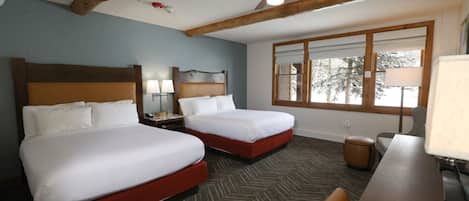 A hotel room with two beds, a wooden ceiling, a large window, and a desk with a lamp.